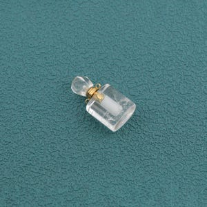 May include: A small, clear, rectangular glass bottle pendant featuring a faceted top and a gold-toned metal collar. The bottle has a white stopper insert and includes a small gold-toned metal loop at the neck for attachment to a chain or cord.