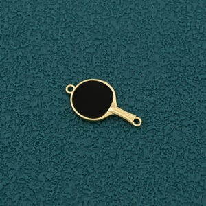 Pode incluir: Um pingente dourado em forma de raquete de pingue-pongue com um centro preto. O pingente tem um pequeno la&ccedil;o na parte superior e inferior da al&ccedil;a para prender a um colar ou pulseira. O pingente est&aacute; em um fundo texturizado azul-petr&oacute;leo.