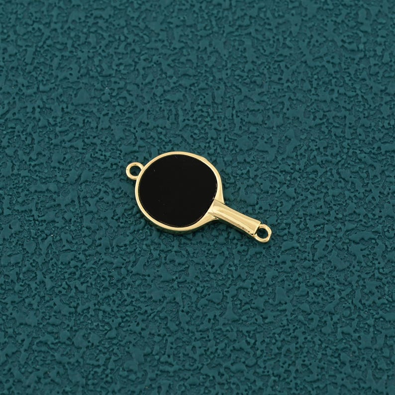 Pode incluir: Um pingente dourado em forma de raquete de pingue-pongue com um centro preto. O pingente tem um pequeno la&ccedil;o na parte superior e inferior da al&ccedil;a para prender a um colar ou pulseira. O pingente est&aacute; em um fundo texturizado azul-petr&oacute;leo.