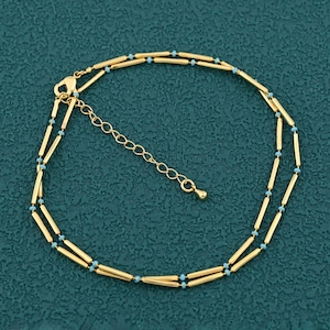 May include: A gold-colored anklet with a double-strand design. The anklet features small, light blue beads interspersed along the length of the gold bars. The anklet has an adjustable chain for a custom fit.