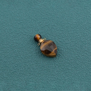 May include: A teardrop-shaped tiger's eye gemstone pendant featuring a faceted surface with brown and gold bands. The pendant includes a small, round tiger's eye stopper attached to a gold-toned metal cap with two tiny loops for hanging or attachment.