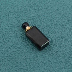 Pode incluir: Um frasco de perfume retangular de vidro preto com uma tampa esférica preta facetada e um colar de metal dourado com dois pequenos anéis circulares nas laterais para fixação, apresentando uma aparência elegante e polida.