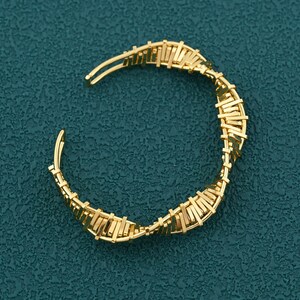 May include: A gold-colored cuff bracelet with a woven design. The bracelet is open at the ends and features a series of small, rectangular bars arranged in a spiral pattern. The background is a textured teal color.