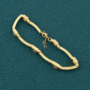 Può includere: Un braccialetto dorato dal design delicato. Il braccialetto presenta accenti rettangolari e una chiusura sicura. Il gioiello è esposto su una superficie color verde acqua. Questo elegante braccialetto è adatto a varie occasioni.