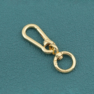 May include: A gold-toned metal lobster claw clasp featuring an elongated, rectangular-shaped hook with a spring-loaded gate, connected to a circular swivel ring. The hardware has a smooth, polished finish and a reflective surface throughout.