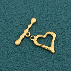 May include: A gold-toned toggle clasp set consisting of a T-bar component and an open heart-shaped ring component. Both pieces feature a textured, irregular surface finish, providing a distinct appearance for jewelry assembly and closure.