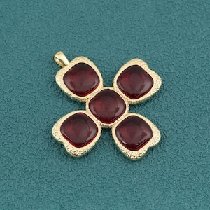 May include: A gold-toned metal pendant featuring five square-shaped, deep red glass cabochons arranged in a cross pattern. The metal frame has a textured, granular surface finish and includes a small loop at the top for attachment to a chain.