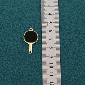 Pode incluir: Um berloque dourado em forma de raquete de pingue-pongue com um centro preto. O berloque tem um pequeno la&ccedil;o na parte superior para prender a uma corrente ou pulseira. Uma r&eacute;gua est&aacute; ao lado do berloque.