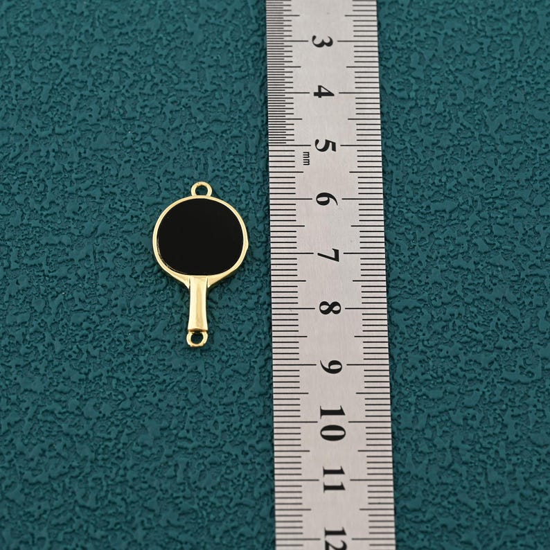 Pode incluir: Um berloque dourado em forma de raquete de pingue-pongue com um centro preto. O berloque tem um pequeno la&ccedil;o na parte superior para prender a uma corrente ou pulseira. Uma r&eacute;gua est&aacute; ao lado do berloque.