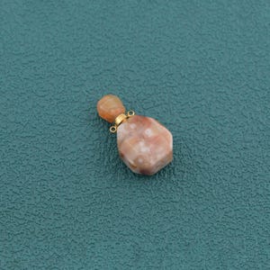 May include: A small, faceted, peach-colored stone pendant with a mottled, semi-translucent appearance. The stone is attached to a gold-toned metal cap featuring a small circular loop at the top for hanging, showcasing a smooth, polished finish.