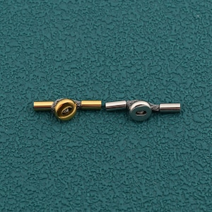 May include: Two toggle clasps for jewelry making, one in a polished gold tone and one in a polished silver tone. Each clasp consists of a cylindrical bar and a circular ring component connected by a short length of braided gray cord material.