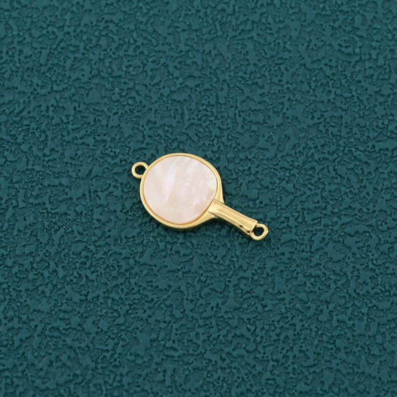 Pode incluir: Um berloque dourado em forma de raquete de pingue-pongue. A face da raquete &eacute; branca perolada clara, e a al&ccedil;a tem um pequeno la&ccedil;o na extremidade. Projetado para fazer joias ou artesanato.