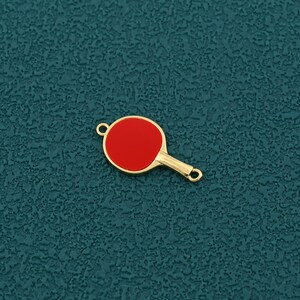 Pode incluir: Um pingente dourado em forma de raquete de pingue-pongue com um centro vermelho. O pingente tem um pequeno la&ccedil;o na parte superior e inferior da al&ccedil;a para prender a um colar ou pulseira. O pingente &eacute; uma vers&atilde;o em miniatura de uma raquete de pingue-pongue.