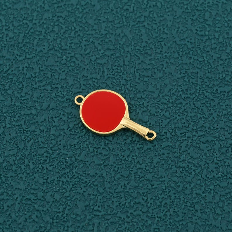 Pode incluir: Um pingente dourado em forma de raquete de pingue-pongue com um centro vermelho. O pingente tem um pequeno la&ccedil;o na parte superior e inferior da al&ccedil;a para prender a um colar ou pulseira. O pingente &eacute; uma vers&atilde;o em miniatura de uma raquete de pingue-pongue.