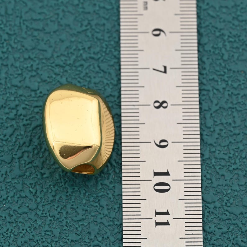 May include: A gold-colored, irregularly shaped bead. The bead has a smooth, polished surface and appears to be made of metal. The bead is approximately 0.75 inches in length. The background is a teal color.