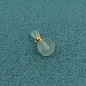 May include: A small, translucent, pale green perfume bottle pendant with a faceted, hexagonal body. The neck of the bottle is capped with a metallic gold-toned collar featuring a small, circular loop attached to the top for hanging purposes.