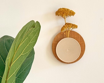 May include: A wall-mounted decorative piece featuring a wooden base with a circular design. A beige, textured, circular element is centered on the wood, with dried yellow flowers. The piece is mounted on a white wall, with large green leaves in the foreground.