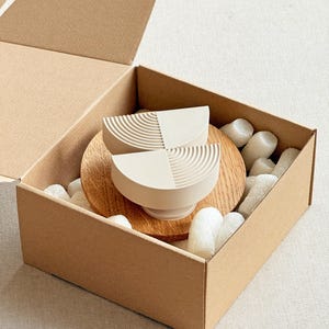 May include: Two off-white, semi-circular bowls with a textured design, presented in a cardboard box with packing material. The bowls sit on a wooden base. The box is open, revealing the contents.