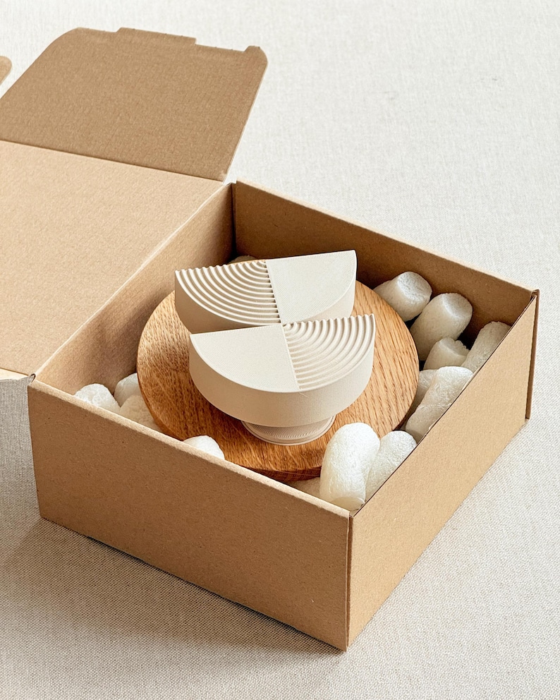 May include: Two off-white, semi-circular bowls with a textured design, presented in a cardboard box with packing material. The bowls sit on a wooden base. The box is open, revealing the contents.