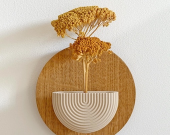 May include: A wall-mounted decorative piece featuring a round wooden base with a beige, semi-circular vase. The vase holds dried yellow flowers. The design is minimalist and modern, suitable for home decor.
