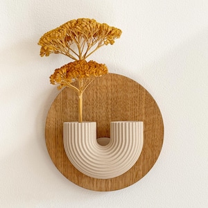Pode incluir: Uma peça decorativa de parede com um vaso em forma de U bege claro, com textura canelada, montado numa base de madeira redonda. O vaso contém flores amarelas secas. Para decoração de casa.