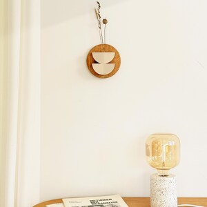 May include: A wooden wall hanging with a circular brown base and two beige, semi-circular cutouts. Dried plants are attached to the top. A table lamp with a white speckled base and a large, amber-colored bulb sits on a wooden table. Books are also on the table.