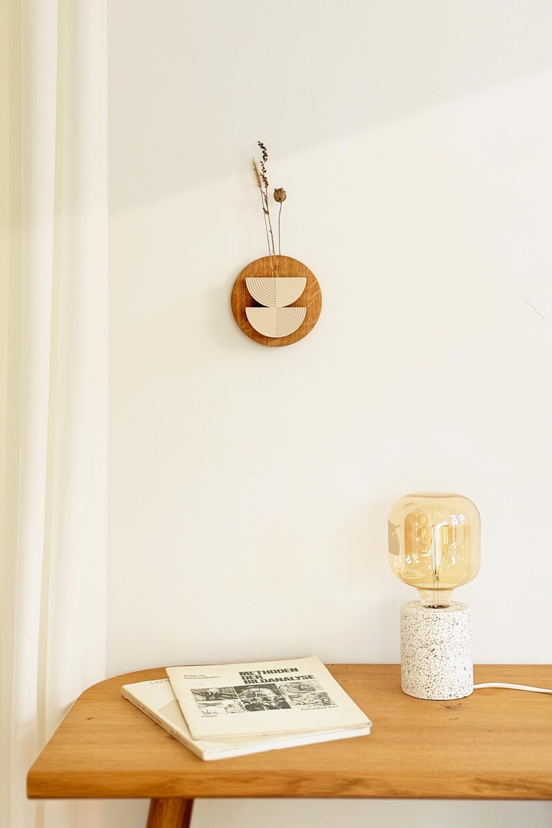 May include: A wooden wall hanging with a circular brown base and two beige, semi-circular cutouts. Dried plants are attached to the top. A table lamp with a white speckled base and a large, amber-colored bulb sits on a wooden table. Books are also on the table.