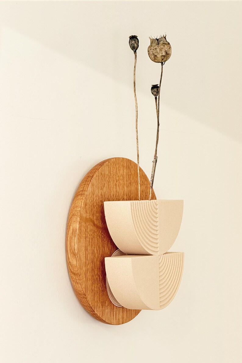 May include: A modern wall-mounted sculpture featuring a light wood oval base with two stacked, cream-colored, semi-circular bowls. Dried seed pods extend upwards from the bowls, creating a minimalist, natural aesthetic.