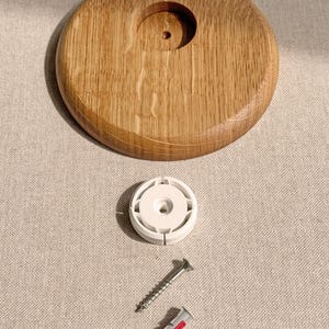 May include: A wooden, circular object with a central hole, accompanied by a white plastic piece, a silver screw, and a red and white wall anchor. The wood has a natural grain pattern.