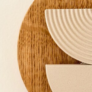 May include: Close-up of a wooden wall art piece. The artwork features a large, circular wooden base with vertical grain. Two off-white, semi-circular elements are layered on top, one with a ridged texture. The piece is set against a neutral background.