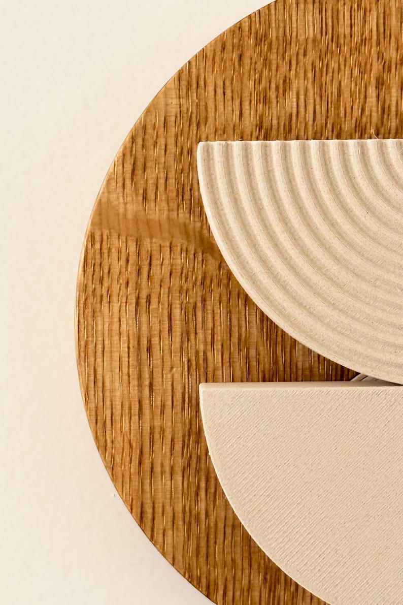 May include: Close-up of a wooden wall art piece. The artwork features a large, circular wooden base with vertical grain. Two off-white, semi-circular elements are layered on top, one with a ridged texture. The piece is set against a neutral background.