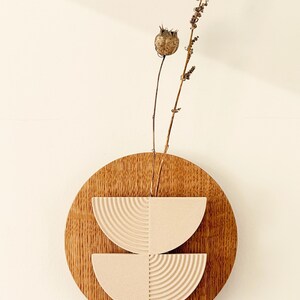 May include: A wooden wall art piece featuring a circular wooden base with two semi-circular, textured, off-white elements. Dried plant stems and seed pods extend from the center. The artwork is mounted on a light-colored wall.