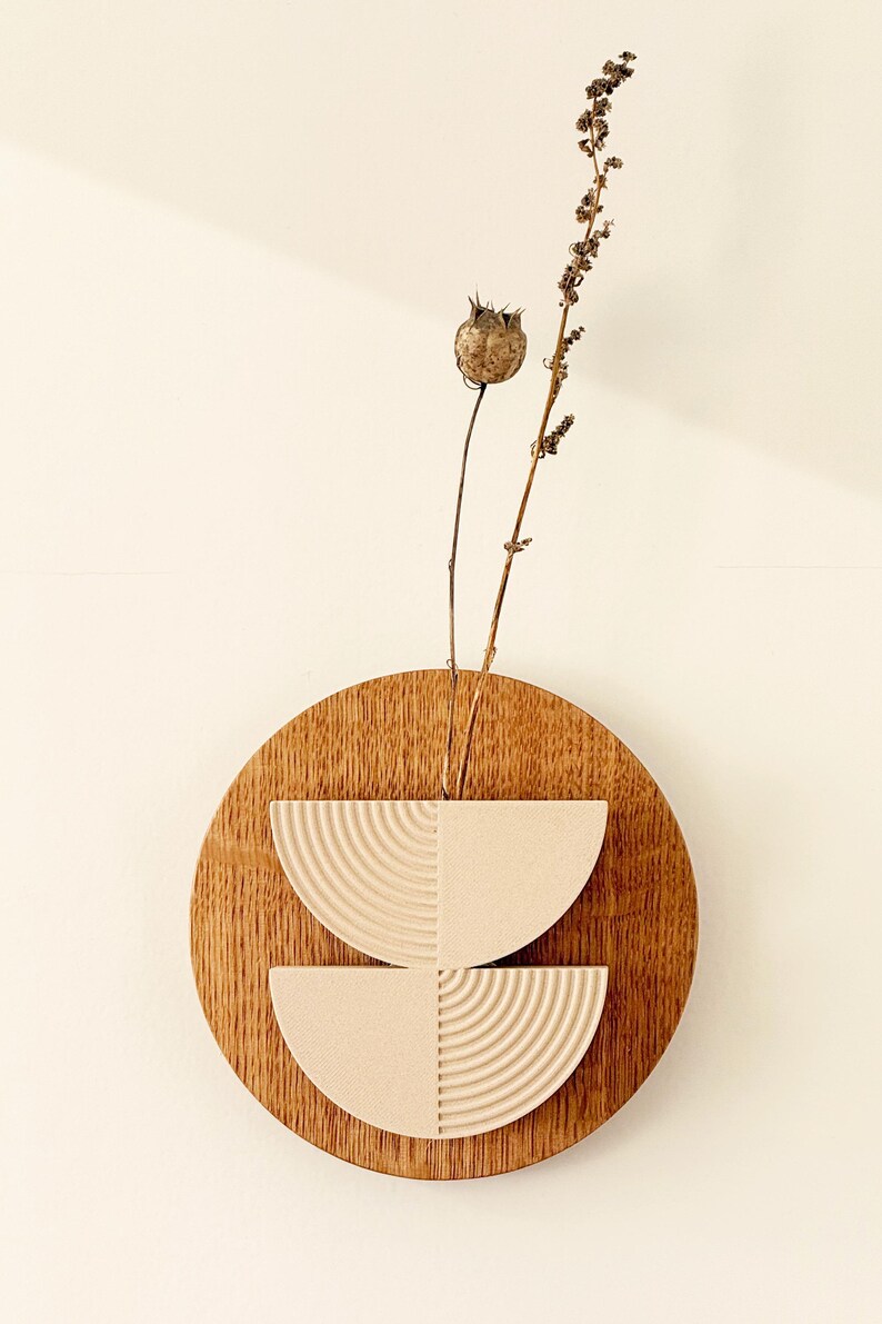 May include: A wooden wall art piece featuring a circular wooden base with two semi-circular, textured, off-white elements. Dried plant stems and seed pods extend from the center. The artwork is mounted on a light-colored wall.
