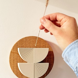 May include: A round, wooden wall art piece with a geometric design. The art features two semi-circular, textured, off-white shapes on a brown wooden base. A hand holds a dried plant stem near the art, adding a natural element.