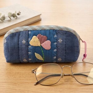 May include: A blue denim eyeglasses case with a floral applique in yellow, pink, and red gingham. The case has a zipper closure with a pink pull cord. A pair of round-framed eyeglasses rests on a wooden surface.