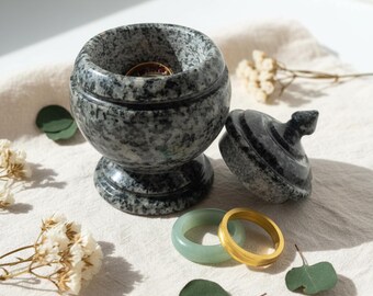 May include: A gray speckled stone jewelry box with a lid, holding rings. The box is round with a pedestal base. Two rings are visible outside the box: a gold band and a light green ring. Dried flowers and leaves are scattered around.