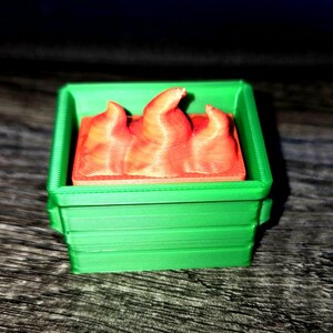 May include: A 3D-printed miniature green dumpster with a red flame design inside. The dumpster has a rectangular shape with horizontal lines on the sides. The flames are orange and have a textured appearance.