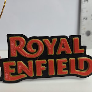 Puede incluir: Un logotipo de Royal Enfield negro y rojo, posiblemente un llavero o un amuleto. El logotipo presenta las palabras "ROYAL ENFIELD" en una fuente audaz y estilizada con un contorno dorado. Una cuerda dorada está unida en la parte superior izquierda.