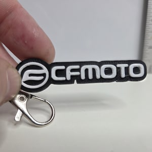 May include: Black and white keychain with the text "CFMOTO". The keychain has a silver clasp. A ruler is in the background.