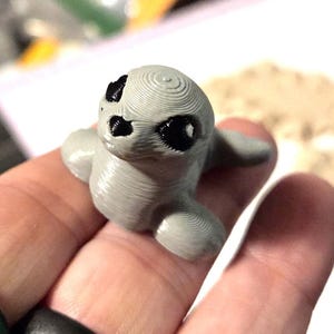 May include: A small, gray 3D-printed seal figurine with black eyes and a textured surface. The seal has flippers and is lying down. The figurine is held in a hand.
