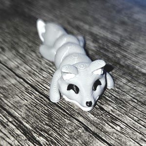 Puede incluir: Figura de zorro gris claro impresa en 3D, tumbada. El zorro tiene ojos negros y nariz negra, con detalles blancos en las orejas y la cola. La figura descansa sobre una superficie de madera.