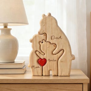 Op de afbeelding: Houten familieberenpuzzel met de woorden "Papa", "Mama" en "Anita" gegraveerd. Een rood hart staat in het midden. De puzzel staat op een houten nachtkastje naast een lamp met een beige lampenkap.