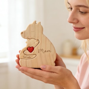 Op de afbeelding: Een houten beeldje in de vorm van een moeder en kind, met een rood hart en het woord "Mom" gegraveerd. De naam "Sarah" is ook gegraveerd. Het beeldje wordt in de handen van een persoon gehouden.