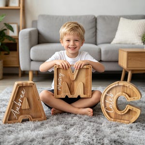 Può includere: Tre salvadanai in legno a forma di lettere A, M e C. Ogni salvadanaio presenta un pannello frontale trasparente ed è personalizzato con una scritta bianca in corsivo che riporta Aubree, Maisey e Caleb sui telai in legno.