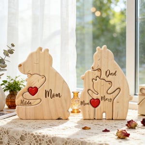 Op de afbeelding: Twee houten berenfamiliefiguren. E&eacute;n toont een beer die een kleinere beer omhelst met een rood hart, het woord "Mom" en de naam "Hilda". De andere heeft drie beren met de woorden "Dad", "Mom" en "Mary".