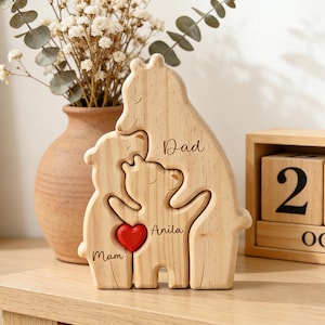 Op de afbeelding: Houten familie berenbeeldje met de woorden "Dad", "Anila" en "Mom" gegraveerd. Een rood hart staat in het midden. Een vaas met gedroogde bloemen en een houten blokkalender staan op de achtergrond. De kalender toont de datum 25 oktober.