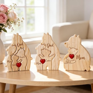 Op de afbeelding: Drie houten berenfamiliefiguren met gegraveerde namen en een rood hart. De beren zijn lichtbruin en staan op een houten tafel. Een vaas met roze en witte rozen staat in de buurt.