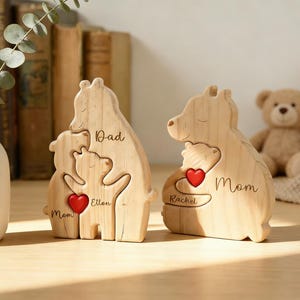 Op de afbeelding: Twee houten berenfamiliefiguren. E&eacute;n heeft "Papa", "Mama" en "Ellen" met een rood hart. De andere heeft "Mama", "Rachel" en een rood hart. De figuren staan op een houten oppervlak.