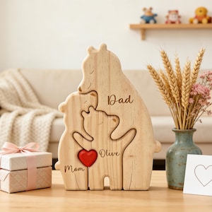 Op de afbeelding: Houten puzzel van een berenfamilie met de woorden "Papa", "Mama" en "Olive" gegraveerd. Een rood hart staat in het midden. Een geschenkdoos met een roze lint en een vaas met tarwe en bloemen zijn ook aanwezig.