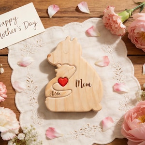 Op de afbeelding: Een houten puzzel in de vorm van een beer met een rood hart, de naam "Hilda" en het woord "Mom" gegraveerd. De puzzel ligt op een witte doily, omringd door roze bloemen, bloemblaadjes en een kaart met "Happy Mother's Day".